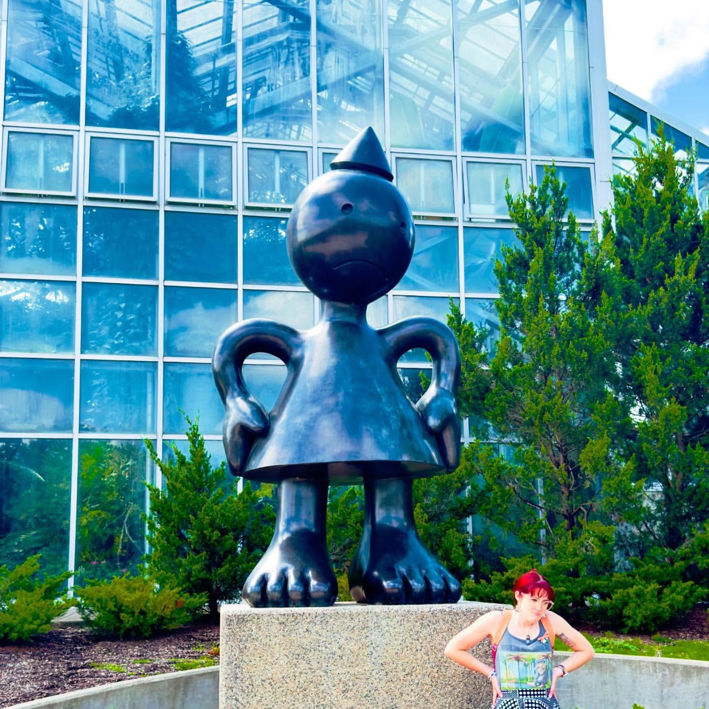 A young woman with red hair posing with her arms up on her hips, looking down and to the right, mirroring a tall sculpture titled "Mad Mom"